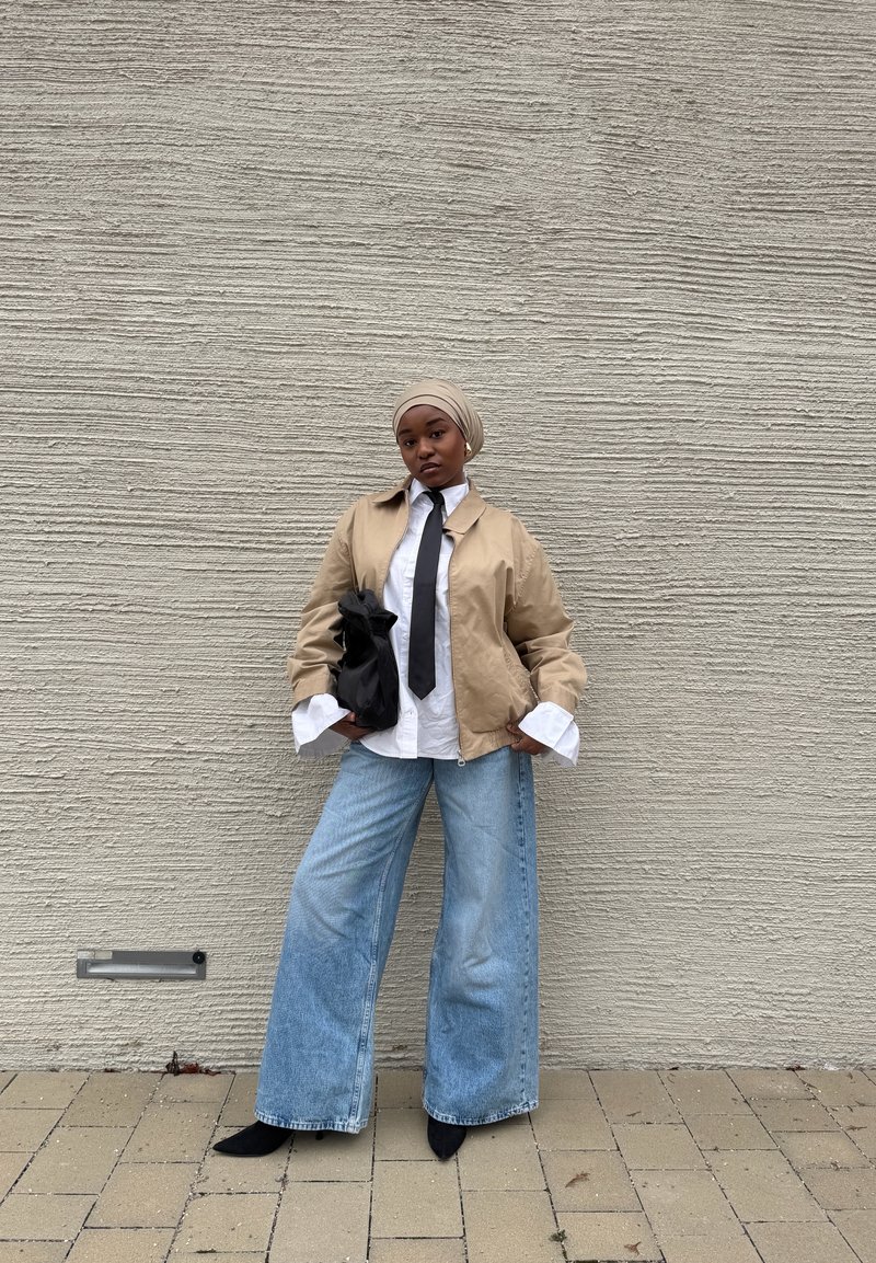 Γυναίκα με μπεζ σακάκι, λευκό πουκάμισο με φαρδιές μανσέτες, μαύρη γραβάτα, ανοιχτό μπλε τζιν παντελόνι με φαρδιά μπατζάκια, μαύρα παπούτσια και μπεζ μαντήλι στο κεφάλι, στέκεται μπροστά σε υφή τοίχου.