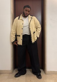 Bărbat cu părul împletit, purtând o jachetă crem cu detalii negre, cămașă albă, pantaloni negri cu croială largă și pantofi negri, stând în interior.