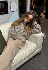 Beige denimjacka och byxor, oversize-passform, med lätta texturer. Bruntonade solglasögon. Sittande på en vit lädersoffa. Grön kudde.