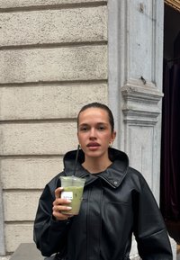Osoba drži prozirnu čašu zelene napitke sa slamkom, obučena u crnu kožnu jaknu s visokim ovratnikom i glatkom teksturom.