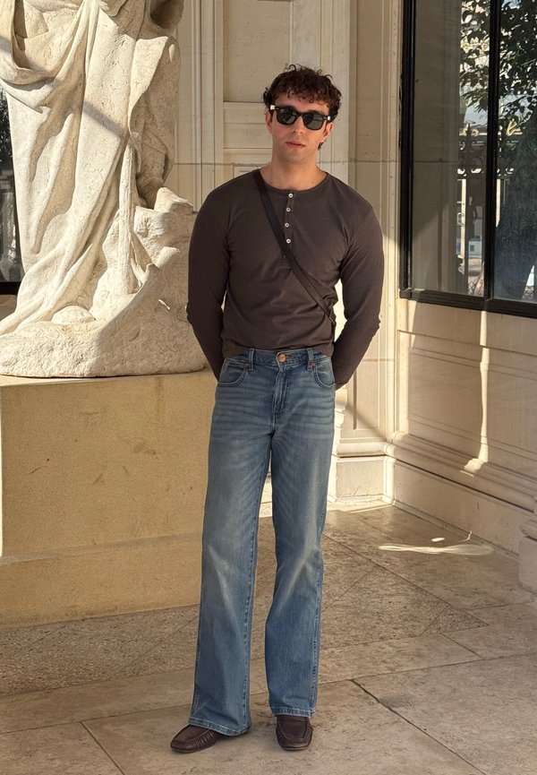 Man wearing dark long-sleeve shirt, blue jeans, sunglasses, and crossbody bag standing indoors near a large stone sculpture.