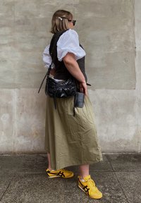 Μαύρο γιλέκο πάνω από μια λευκή μπλούζα με φουσκωτά μανίκια, ελιά-πράσινη φούστα και κίτρινες αθλητικές παπούτσια με μαύρες λεπτομέρειες. Μαύρη τσάντα με φερμουάρ.