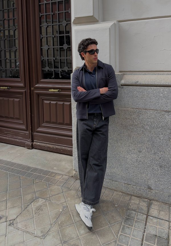 Homme portant une veste sombre, une chemise bleu marine, un jean noir à jambes larges, des baskets blanches et des lunettes de soleil, appuyé contre un mur de pierre gris près de portes en bois.