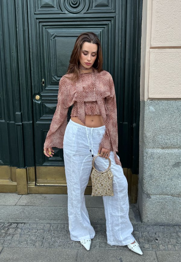 Kvinde iført mønstret brunt crop top og hvide vidde bukser holder en lille perlet håndtaske og står foran en mørkegrøn dør med sten- og messingdetaljer.