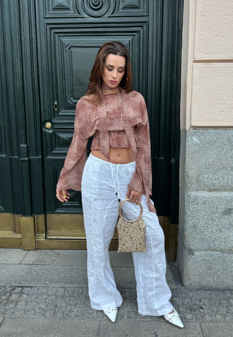 Γυναίκα με καφέ crop top με σχέδια και λευκό φαρδύ παντελόνι κρατά μια μικρή τσάντα με χάντρες, στέκεται μπροστά σε μια σκούρα πράσινη πόρτα με πέτρινες και ορειχάλκινες λεπτομέρειες.