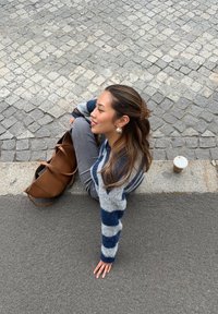 Grauer Pullover mit marineblauen Streifen, graue Hose, braune Tasche und ein To-go-Kaffeebecher. Person sitzt auf dem Gehweg zwischen Pflastersteinen.