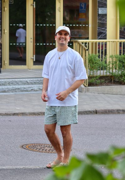 Camiseta blanca de gran tamaño, pantalones cortos verdes con patrón, sandalias beige; de pie al aire libre cerca de puertas de cristal con vegetación de fondo.