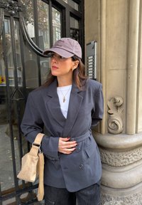 Femme en blazer gris et casquette de baseball, debout près d'une colonne de pierre ornée et d'une grille en métal noir, tenant un sac à bandoulière beige.