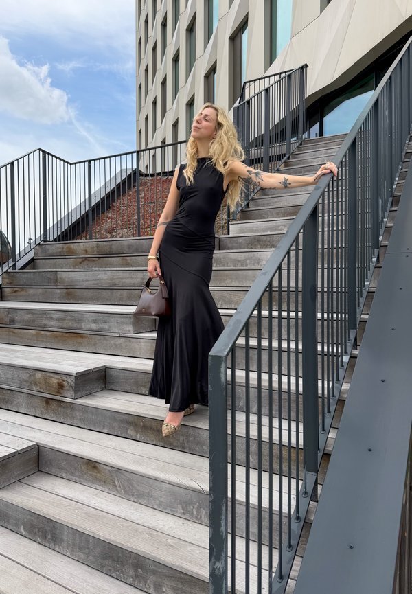 Femme en longue robe noire debout sur un escalier en bois extérieur, tenant la rampe et un sac à main, bâtiment moderne en arrière-plan sous un ciel partiellement nuageux.