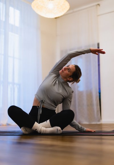 Una persona practica yoga en una esterilla, vestida con una camiseta de manga larga gris claro y leggings negros. El suelo es de madera, y una luz suave ilumina el espacio.