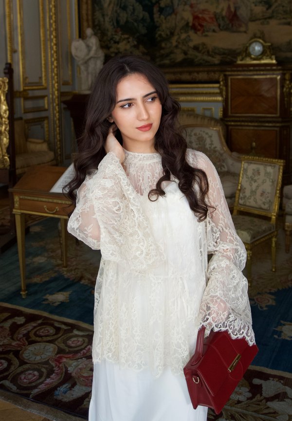 Jeune femme aux longs cheveux foncés portant une robe en dentelle blanche tient une pochette rouge, debout dans une pièce élégante avec des meubles ornés et un tapis à motifs.