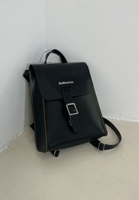 Black leather backpack featuring a flap closure, silver buckle accent, yellow stitching, and adjustable straps. It has a smooth texture with minimal detailing.