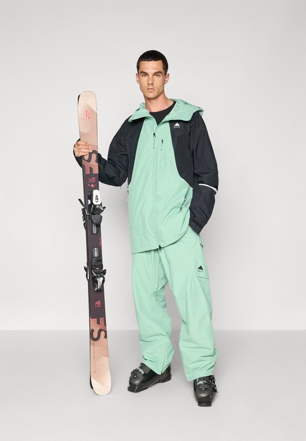 Traje de esquí en verde menta y negro, con chaqueta con cremallera y pantalones. El modelo sostiene esquís oscuros con fijaciones, vistiendo botas de esquí negras.