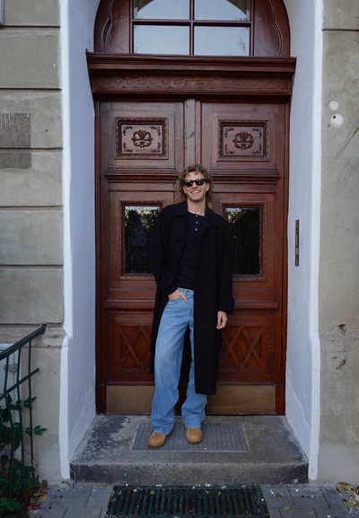Abrigo largo negro, camisa oscura, vaqueros azul claro y zuecos color canela. De pie frente a unas elegantes puertas dobles de madera con paneles decorativos.