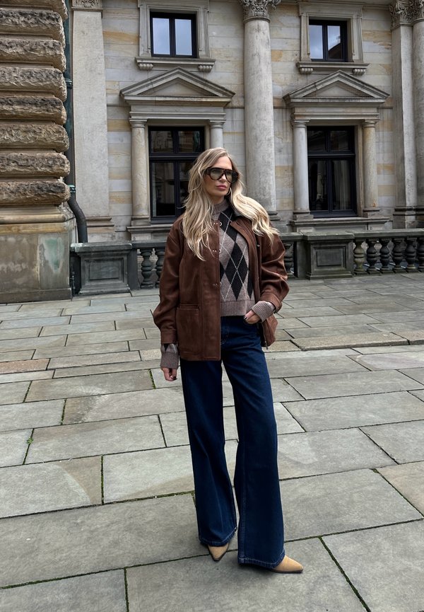 Giacca di pelle marrone su un maglione a fantasia marrone e beige, abbinata a jeans svasati blu scuro e décolleté beige a punta.
