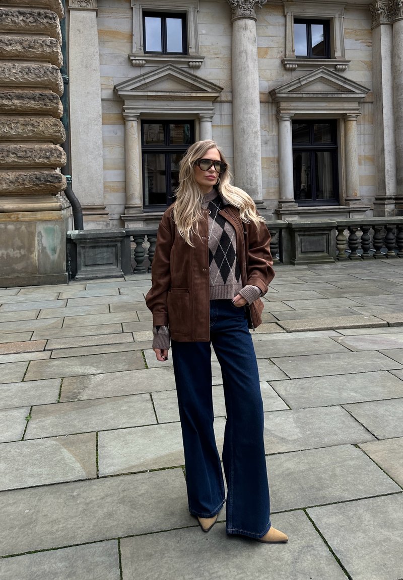 Brown leather jacket over a patterned brown and beige sweater, paired with flared dark blue jeans and beige pointed heels.