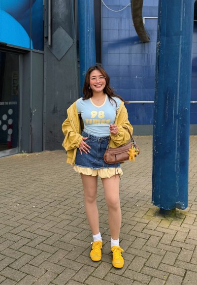 Joven mujer con gafas, chaqueta amarilla, camiseta azul con el texto "98 TIMES", falda vaquera y zapatillas amarillas, de pie en una calle pavimentada con un edificio azul de fondo.
