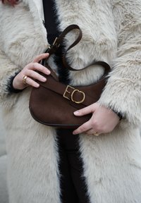 Άτομο με μπεζ παλτό από συνθετική γούνα κρατάει μικρή καφέ σουέντ τσάντα με χρυσή αγκράφα, φοράει χρυσά δαχτυλίδια στα δάχτυλα.