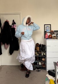 Άτομο που φοράει ανοιχτό μπλε μπουφάν με κουκούλα με γούνινη επένδυση και λευκή ροή φούστα, ποζάροντας μέσα σε ένα δωμάτιο με παπούτσια και μια συρταριέρα κοντά.