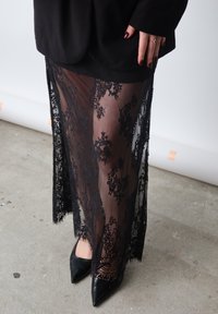 Gonna nera in pizzo con motivi floreali, design trasparente e spacco. Abbinata a tacchi neri a punta su un pavimento in cemento.