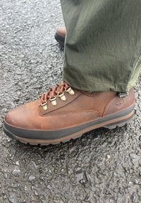 Barna bőr túracipő fém szemekkel, piros fűzővel, gumitalppal és texturált felülettel. Zöld nadrággal viselve aszfalton.