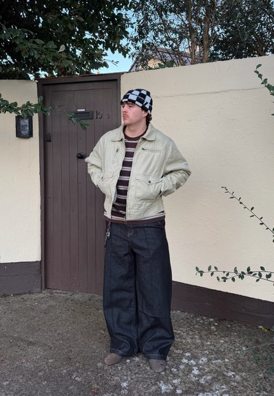 Persona con un sombrero de cuadros, chaqueta beige, camiseta a rayas y jeans anchos oscuros, de pie con las manos en los bolsillos junto a una puerta marrón y una pared crema.