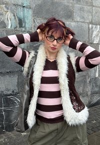 Jeune femme ajustant ses cheveux avec les mains, portant des lunettes rondes, un pull rayé rose et marron, une veste en fourrure, et un pantalon vert olive contre un mur en pierre.