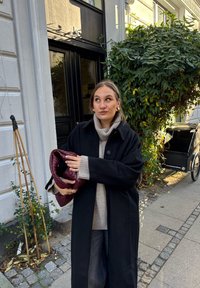 Čierny kabát cez sivý sveter, drží bordovú tkanú tašku. V pozadí je rastlina s listami a bicyklový vozík na kamennej ceste.