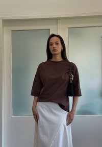 T-shirt marrone, dal taglio morbido e con maniche corte, abbinato a una gonna avvolgente bianca. Gli accessori includono una piccola borsa nera con una tracolla intrecciata.