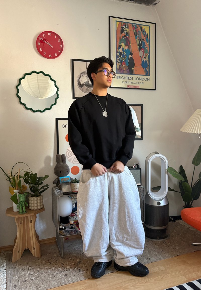 Jeune homme portant un pantalon gris trop grand et un pull noir, debout dans une pièce décorée avec des plantes, une lampe, une horloge rouge et des œuvres murales colorées.