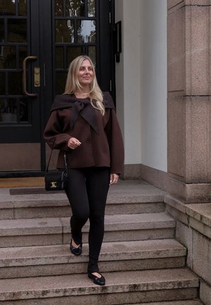 Suéter de lana marrón con cuello ancho y detalle de lazo, combinado con leggings negros y zapatos planos, sosteniendo un pequeño bolso negro. Pasos en el fondo.