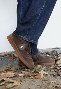 Hnedé semišové tenisky s gumovou podrážkou, zdobené zlatým emblémom, viazaním na šnúrky a jemnými textúrnymi detailmi, v kombinácii s modrými džínsami.