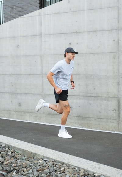 Hombre corriendo por un sendero, vistiendo una camiseta deportiva gris claro, pantalones cortos negros, zapatillas blancas y una gorra oscura. Alrededor hay una pared de concreto.