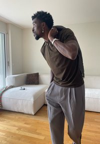 Mężczyzna z krótkimi kręconymi włosami i kolczykiem, ubrany w ciemną koszulę i szare spodnie, trzymający kurtkę na ramieniu, stojący w jasnym salonie.