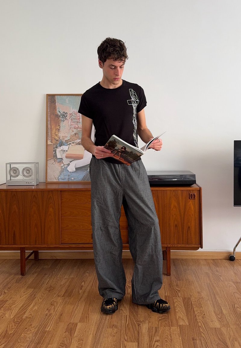 Person wearing a black t-shirt with a graphic design, grey wide-leg trousers, and black shoes with gold accents, reading a magazine indoors.