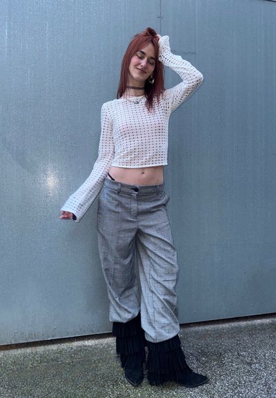 Joven con cabello rojo posando frente a una pared gris, vistiendo un crop top de manga larga de malla blanca, pantalones de cuadros y botas negras con flecos en capas.