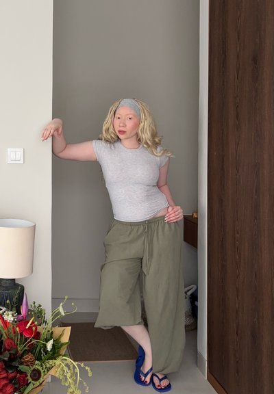 Mujer joven con pelo largo y rubio y diadema se apoya en una pared gris, lleva puesta una camiseta gris, pantalones anchos color oliva y chanclas azules en el interior.