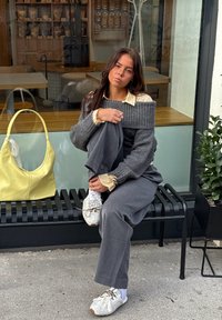 Grauer gerippter Pullover, darunter ein beigefarbener Hemd mit Kragen, graue weite Hose, weiße Sneakers, gelbe Handtasche auf einer schwarzen Bank, Pflanze daneben.