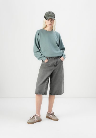 Sudadera de algodón verde claro con un corte holgado, combinada con bermudas de denim gris y zapatillas beige con cordones rosas. Una gorra gris completa el look.