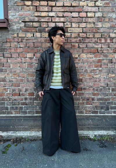 Chaqueta de cuero marrón, suéter a rayas verdes y pantalones de pierna ancha negros. Gafas de sol y fondo de pared de ladrillo realzan la estética urbana.