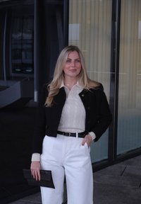 Veste noire raccourcie, pull crème en maille torsadée, pantalon blanc taille haute et pochette noire texturée. Éclairage chaleureux et arrière-plan en verre.