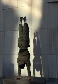Une personne portant un sweat à capuche vert foncé et un pantalon fait un poirier sur un bloc gris contre un mur gris clair, projetant une ombre sur la surface.
