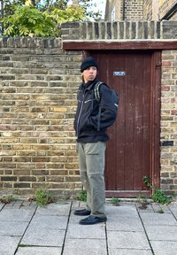 Casaco preto com fecho éclair e detalhes refletivos, calças verdes e sapatos pretos de slip-on. De pé ao lado de uma parede de tijolos e uma porta de madeira.