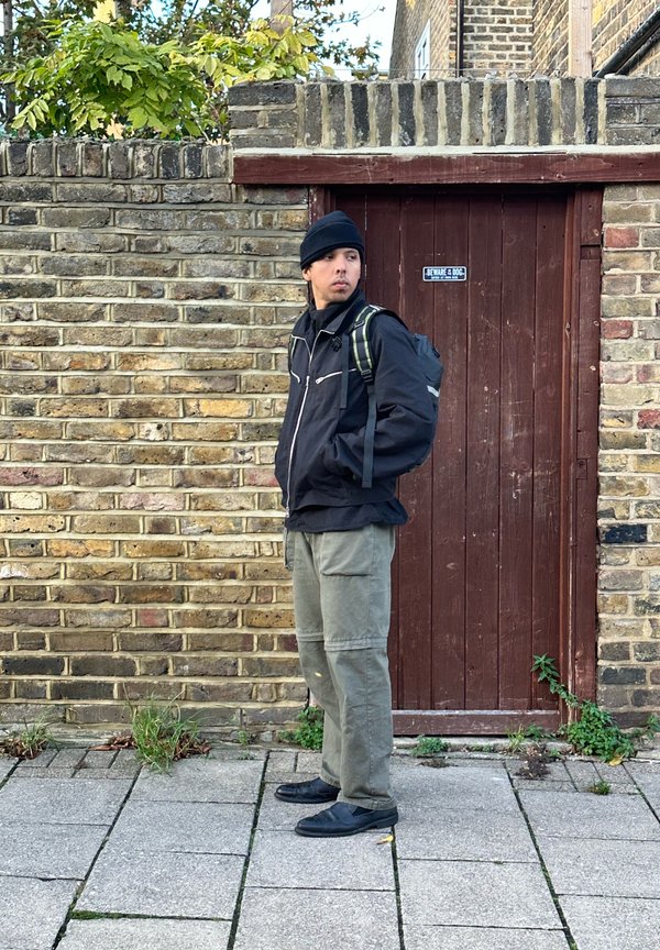 Schwarze Zip-Jacke mit reflektierenden Akzenten, grüne Hose und schwarze Slipper. Steht neben einer Ziegelmauer und einer Holztür.