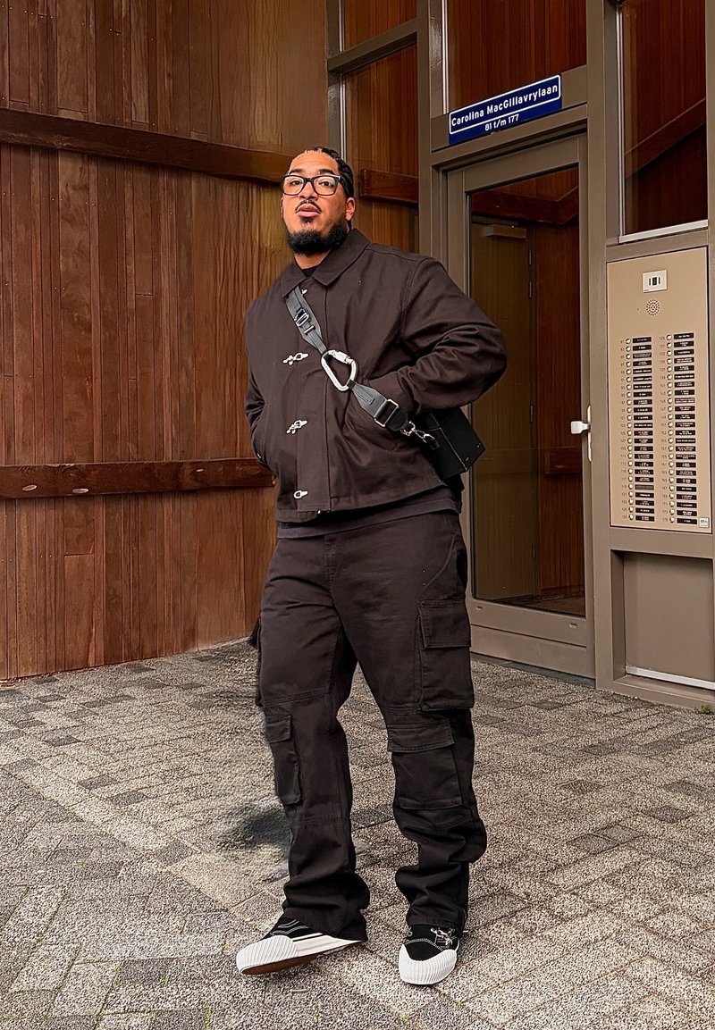 Homme portant une veste noire et un pantalon cargo, debout sur un trottoir pavé à côté de l'entrée d'un bâtiment aux murs en bois et avec un panneau d'interphone.