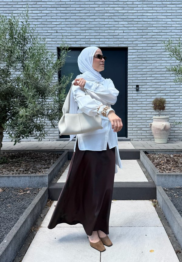 Femme portant un hijab blanc, un chemisier blanc, une jupe sombre, des lunettes de soleil, tenant un sac à main blanc, debout sur un chemin pavé avec des plantes et un mur en briques en arrière-plan.