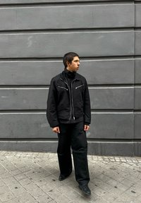Giovane in giacca e pantaloni neri che cammina su un marciapiede piastrellato accanto a un muro di cemento grigio, guardando a destra.