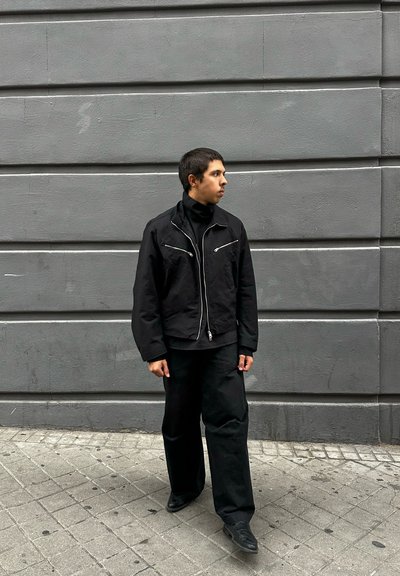 Joven con chaqueta y pantalones negros caminando por una acera de baldosas junto a una pared de concreto gris, mirando hacia la derecha.