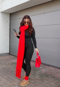 Écharpe rouge surdimensionnée, haut noir ajusté, leggings noirs, sabots marron, chaussettes rouges et petit sac à main rouge. L'arrière-plan présente une porte de garage grise.