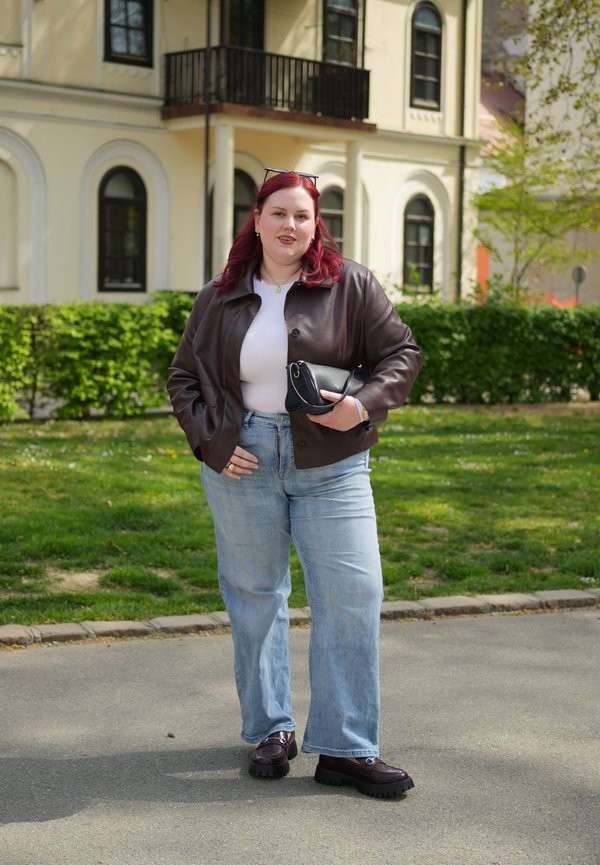 Žena s červenými vlasy má na sobě hnědou koženou bundu, bílý top, modré džíny a černé boty, drží černou kabelku, stojí venku u budovy.